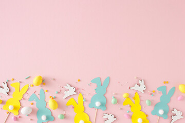 Easter decor concept. Top view photo of yellow white eggs cute rabbits paper bunny toppers and sprinkles on pastel pink background with emty space