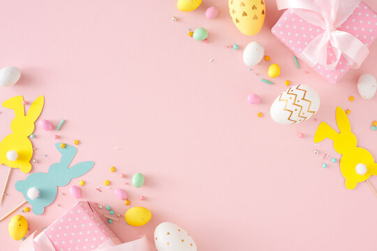 Easter concept. Flat lay photo of colorful eggs gift boxes cute paper bunnies and sprinkles on isolated pastel pink background with copyspace. Holiday card idea