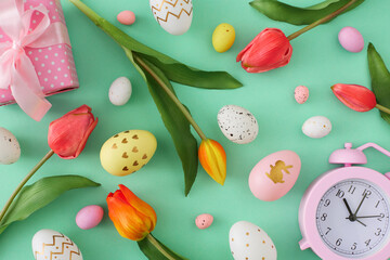 Easter concept. Top view composition of colorful easter eggs tulips flowers gift box and pink alarm clock on isolated turquoise background