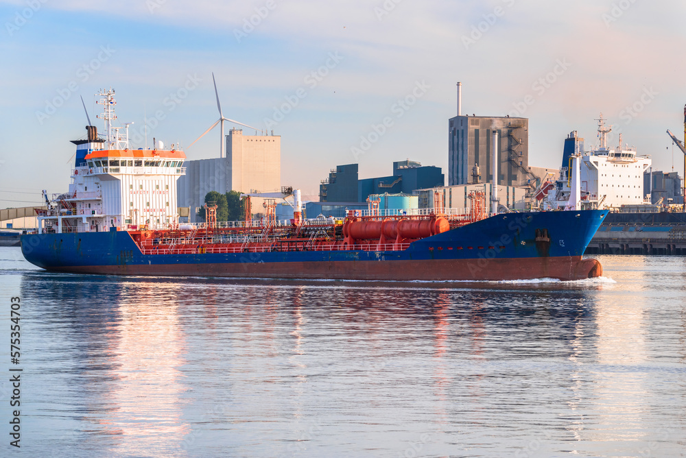 Wall mural chemical tanker leaving a commercial port at sunset. docks, warehouses and wind turbinas are in back