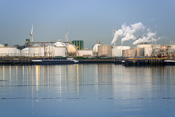 Fototapeta premium Harbourside oil refinery with white smoke belching out the smokestacks at dusk. Wind turbines are in background.