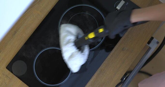 Woman in rubber gloves cleaning black ceramic cooktop with a hot steam cleaner.