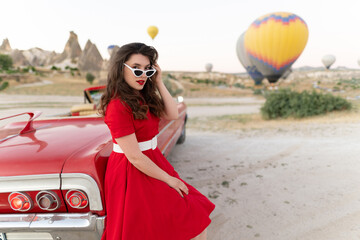 beautiful girl in retro style posing near a vintage red cabriolet car on background of balloons in...