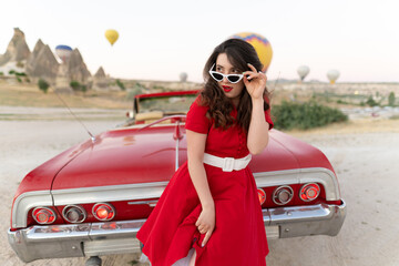 beautiful girl in retro style posing near a vintage red cabriolet car on background of balloons in...