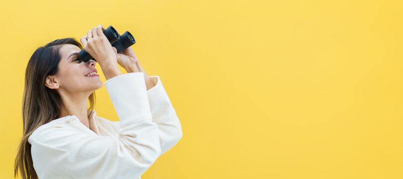 Beautiful caucasian white young woman looking through the twin lens binocular. Woman using binocular portrait on yellow background.