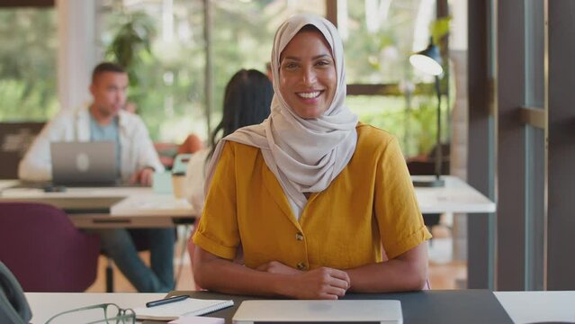 Mature businesswoman wearing headscarf working on laptop at desk in modern office pauses to look out of the window and smile - shot in slow motion