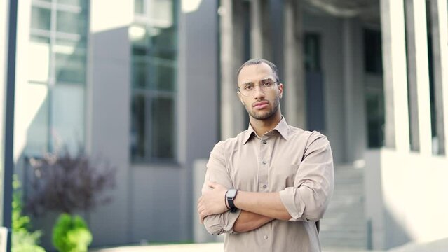 Portrait of a young adult man in a shirt and glasses seriously looking at the camera while standing on the street near an office building. Confident handsome mixed race male posing with crossed arms