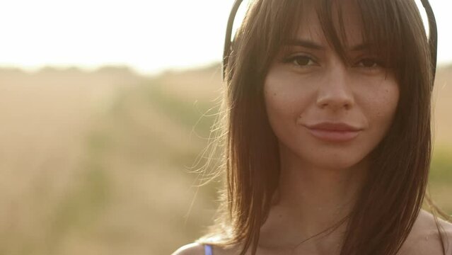 Close-up portrait of a young brunette girl woman cute with a sly seductive look listening to music in headphones while standing in a field outdoors at sunrise