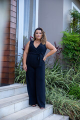 fat woman empowered in front of a house, successful businesswoman with hand on waist wearing elegant black jumpsuit, person in front of a house