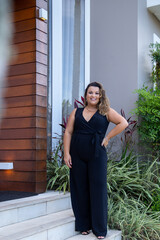 fat woman empowered in front of a house, successful businesswoman with hand on waist wearing elegant black jumpsuit, person in front of a house