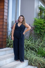 fat woman empowered in front of a house, successful businesswoman with hand on waist wearing elegant black jumpsuit, person in front of a house