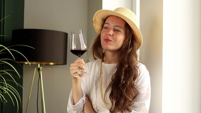 Beautiful young brunette woman drinking red wine from wineglass, enjoying rest time on party in restaurant, wearing white shirt and fashion summer hat. Female sommelier on wine degustation
