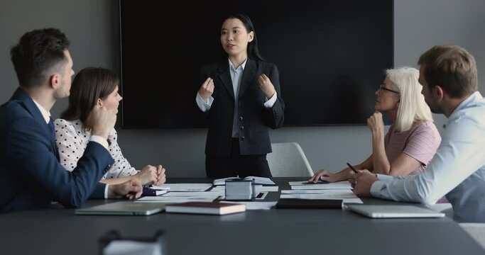 Asian business coach makes presentation, share information to team, looks confident leads formal meeting with company investors or clients sit at desk gathered in boardroom. Corporate training event