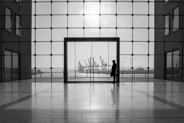 Foto op Plexiglas Blick durch ein großes modernes Glasfenster, mittig eine große Glastür,  Gebäudeteile, im Hintergrund die Silhouetten von Hafenkränen und die Silhouette eines Mannes zu sehen, der am Türrahmen lehnt © Andrea Gaitanides