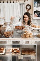 Beautiful young female seller working in a confectionery shop or bakery. She is selling and packing...