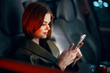 horizontal portrait of a stylish, luxurious woman in a green leather coat, sitting in a black car at night on the passenger seat, holding her phone during a trip illuminated by a red light