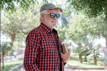 Fototapeta premium Handsome senior bearded man walking in urban park with a book under his arm. Smiling pensioner enjoying free time and retirement