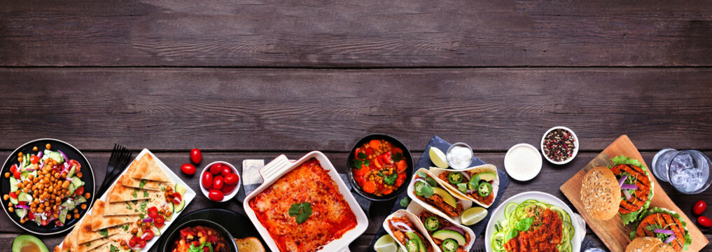 Healthy plant based vegetarian meal bottom border. Top down view on a dark wood banner background. Jackfruit tacos, zucchini lasagna, walnut bolognese zoodles, chickpea burgers, hummus, soups, salad.