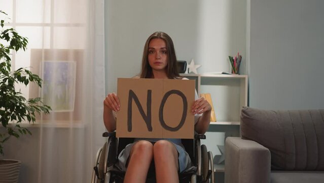 Serious woman with disability shows poster with word No to social inequality looking in camera. Young person with amyotrophic lateral sclerosis sits in wheelchair