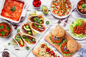 Fototapeta na wymiar Healthy plant based vegetarian meal table scene. Above view on a white wood background. Jackfruit tacos, zucchini lasagna, walnut bolognese zoodles, chickpea burgers, hummus, soups, salad.