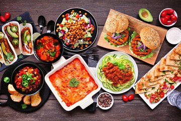 Healthy plant based vegetarian meal table scene. Top view on a dark wood background. Jackfruit tacos, zucchini lasagna, walnut bolognese zoodles, chickpea burgers, hummus, soups, salad.