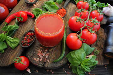 A glass cup with tomato juice on a wooden table is decorated with basil leaves, tomatoes, cherry tomatoes, spices, salt, garlic, hot peppers and peppercorns.
