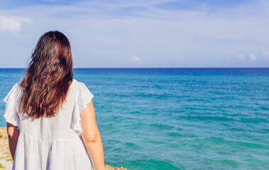 Back view of unrecognizable female tourist with long dark hair wearing white summer dress enjoying picturesque seascape during vacation in Cuba. Anonymous woman admiring waving sea