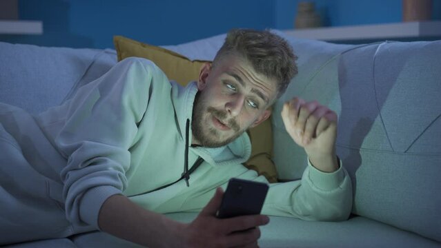 Handsome young man using phone at home in a sad and depressed mood.
Unhappy and lonely young man looking at his phone in a helpless and reluctant mood at home, feeling boring, depressed.
