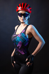 A young female cyclist wearing a safety helmet and glasses, dressed in a bib shorts poses against a black background in the studio.