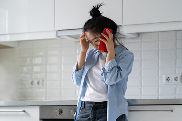 Sad depressed young Asian woman standing at home talking phone, hearing bad news, calling psychologist to receive mental health support, frustrated female making phone call, needs someone to talk to.