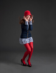 Cute smiling young woman in preppy outfit with short skirt holding down red cloche hat - 575430985