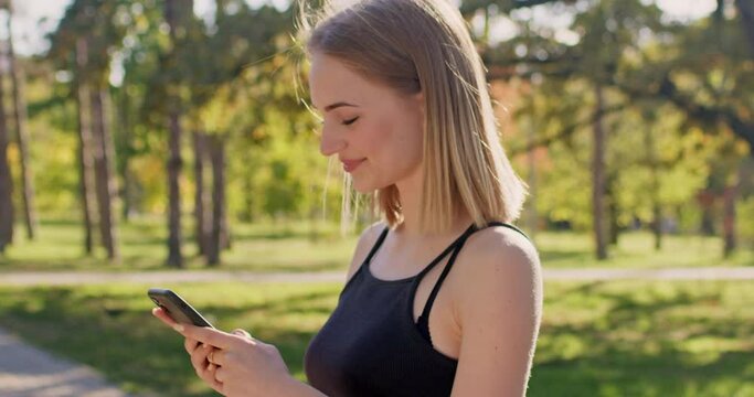 A blonde woman in black sportswear stands in a park with a phone in her hands, smiles and looks at the smartphone screen.