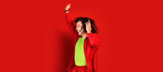 Banner with cute dancing girl in headphones on a bright red background. Curly young woman in red shirt and salad t-shirt. Smiling female listens to music with headphones and jumps