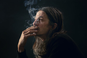 Studio portrait on black background of middle age man with long hair smoking cigarette