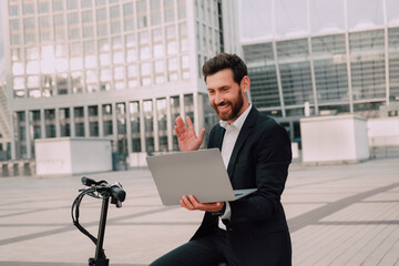 Fototapeta premium modern Successful man in a business suit outdoors near the scooter .Raises his hand up and rejoices in success looking at the laptop