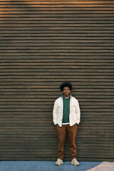 Young cool African American hipster teen guy standing at wooden wall background. Stylish trendy gen z teenage student model looking at camera for authentic portrait outdoors. Vertical full length shot