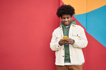 Fototapeta Happy young African American guy standing at color bright red wall outdoors using cell phone, looking at camera holding cellphone enjoying doing online ecommerce shopping in mobile apps, playing game. obraz