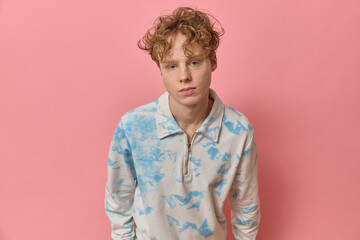 Fototapeta na wymiar Portrait of curly red haired stylish smart teenager with a perfect skin wearing colorful clothing shirt looking at camera posing stands leaning over against pink backdrop copy space in a studio shot.