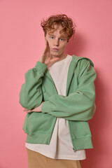 Vertical fashion campaign shot of a young curly haired male with freckles showing duck face wearing trendy colourful clothes standing with folded arms touching his face funny posing in pink studio.