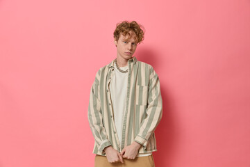 Studio fashion portrait of young confident curly haired ginger man with freckles in trendy clothes green and white striped shirt and chain on neck posing by pink background and looking at camera.