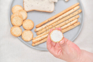 Top view of a hand holding a cracker spread with cream cheese, in the background a plate with wheat bread, crackers and breadsticks. Concepts: gluten and cereals in food.
