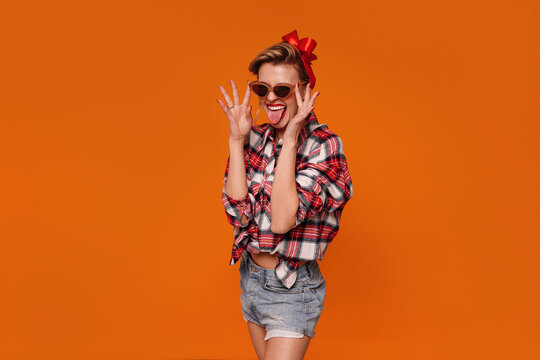 Photo of crazy pinup girl in checkered shirt and short jeans smiling and showing tongue out.