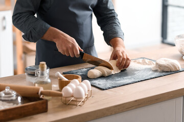 Male baker cutting dough at table in kitchen, closeup