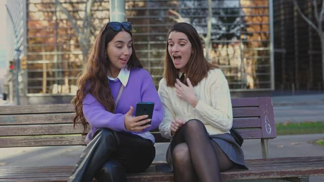 Excited happy young female friends reading unexpected message on mobile phone sitting on bench in city street. Surprised millennial young people receiving good news on smartphone app.