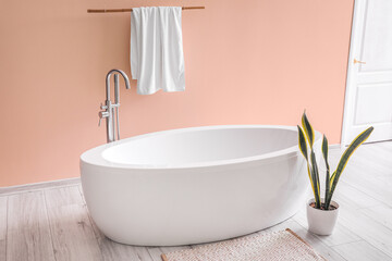 Fototapeta na wymiar Modern bathtub with towel and houseplant near pink wall