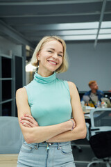 Vertical photo of smiling blonde Caucasian business woman looking at camera standing with arms crossed in office. Executive CEO professional manager posing for business portrait.