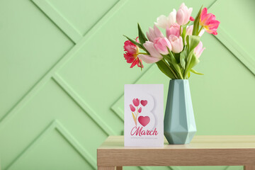 Vase with tulips and greeting card for Women's Day on table near green wall