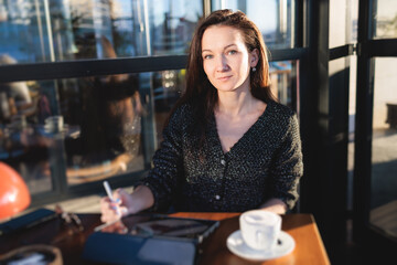 Female young digital artist illustrator drawing digital picture on a graphics tablet with pen stylus in cafe in a sunny day, freelance graphic designer working remotely and sketching with pencil