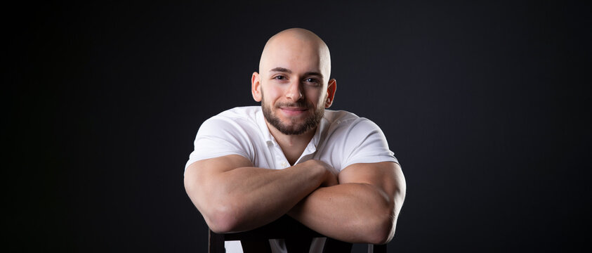turkish student man with bald head and white polo shirt lean on chair back with arms crossed while look at camera and smile with honest and happy face expression