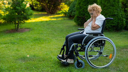 Unhappy elderly woman crying while sitting in a wheelchair on a walk outdoors. 
